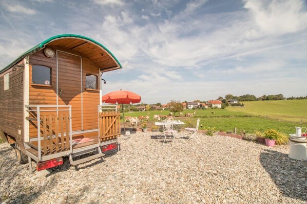 Une cabane en bois avec un toit vert, un parasol rouge et une terrasse en gravier aménagée avec du mobilier d’extérieur.