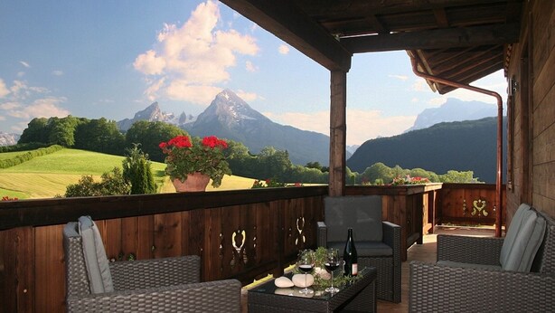 Ein Balkon mit Korbmöbeln, einem Tisch mit Weingläsern und einem Blumentopf mit roten Blüten.