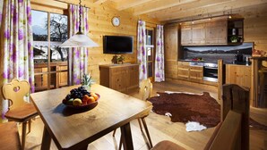 Ein Holzblockhaus-Interieur mit Esstisch, Stühlen, Obstschale, Fernseher, Holzkästen und einer Küchenzeile.