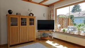 Ein Wohnzimmer mit Holzschrank, Flachbildfernseher, Tisch mit Teller und Fenster mit Blick auf Häuser und Pflanzen.