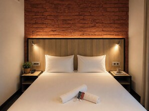 Ein Hotelzimmer mit Backsteinwand, einem Holz-Kopfteil, zwei weißen Kissen, einem ordentlich bezogenen Bett, einem Nachttisch mit Lampe, einer Pflanze und einem zusammengerollten Handtuch mit Toilettenartikeln.
