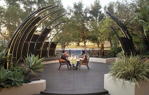 Un área de comedor al aire libre con una mesa circular y sillas, rodeada de plantas y árboles.
