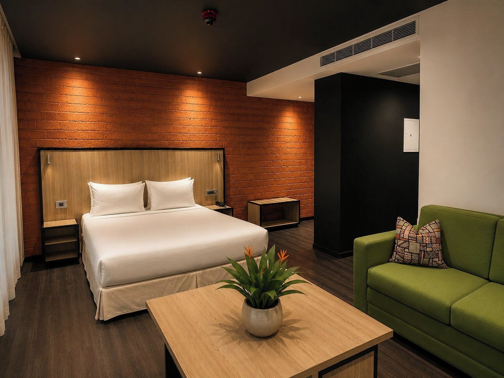A hotel room with a large bed, a green sofa, a small table with a plant, and a brick wall.