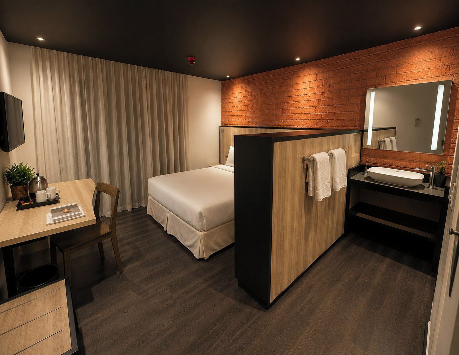 A hotel room with a bed, a desk with a chair, a small table with a plant, a mirror, and a bathroom with a sink and towel rack.