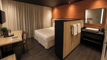Une chambre d’hôtel comprenant un lit, un bureau avec une chaise, une petite table avec une plante, un miroir et une salle de bain avec un lavabo et un porte-serviettes.