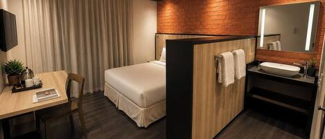 Une chambre d’hôtel comprenant un lit, un bureau avec une chaise, une petite table avec une plante, un miroir et une salle de bain avec un lavabo et un porte-serviettes.