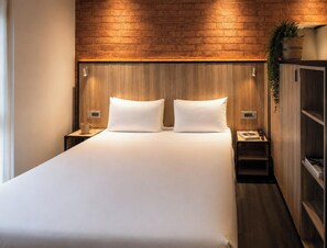 Une chambre d’hôtel avec une tête de lit en bois, un grand lit avec du linge de lit blanc, des tables de chevet avec des lampes et une plante sur la table de chevet.
