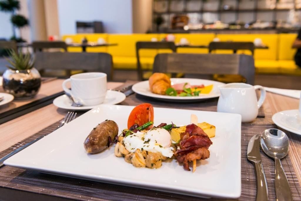 A breakfast setting with a plate of eggs, sausage, and vegetables, a croissant, and a cup of coffee on a table.