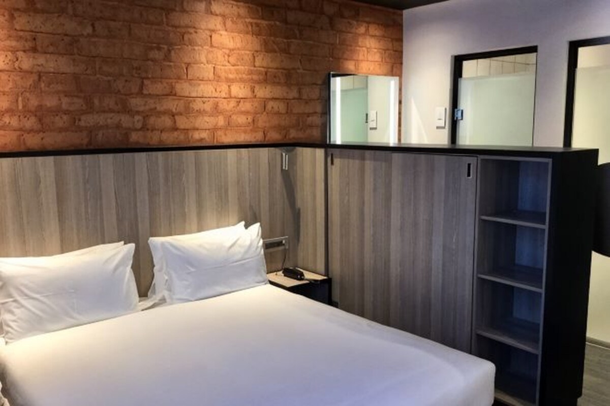 Una habitación de hotel con pared de ladrillo, cama con ropa blanca, cabecero de madera, mesita de noche y armario con puertas espejadas.