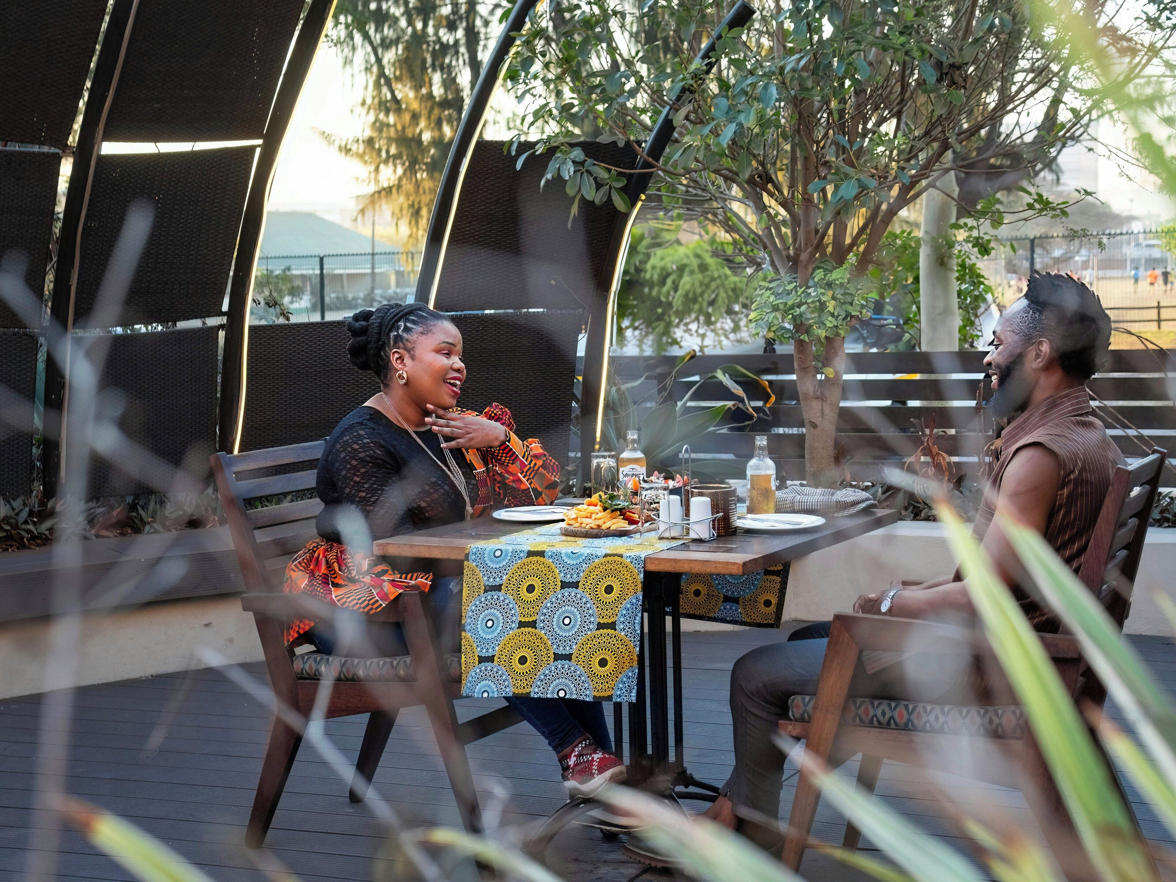 Un espace de restauration en plein air avec une table dressée pour deux personnes, entouré de verdure et délimité par une clôture.