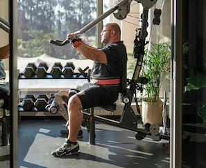 Un homme utilise un appareil de musculation pour les jambes dans une salle de sport, avec des haltères et une plante en arrière-plan.