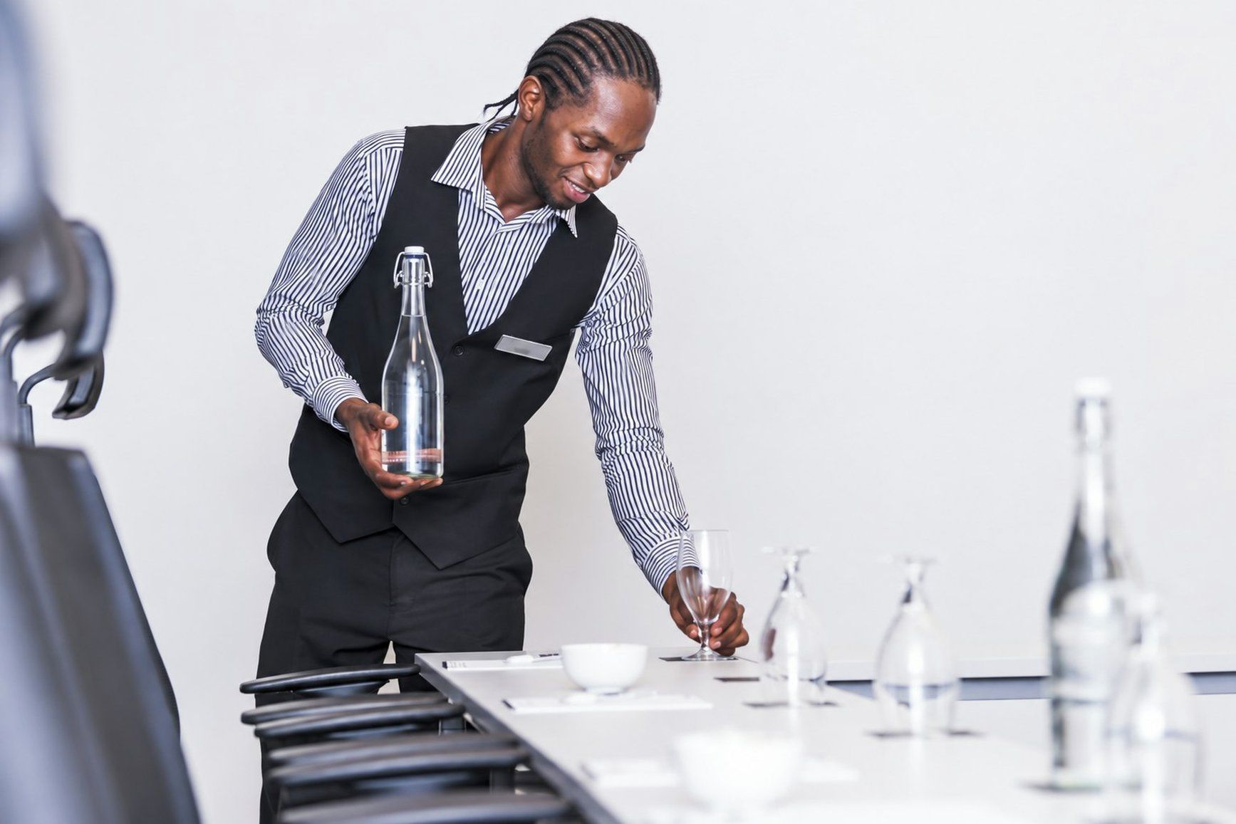 Eine Person in einer schwarzen Weste und gestreiftem Hemd reicht an einen Tisch mit Weinglas, Schüssel und Flasche eine Wasserflasche.