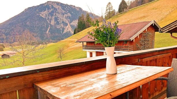 Ein hölzerner Balkon mit einer weißen Blumenvase, mit Blick auf eine Berglandschaft und ein traditionelles Holzhaus.