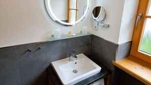 Un baño con un lavamanos blanco, un espejo redondo con luz, una ventana pequeña y un mueble de tocador de madera.