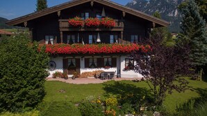 Un chalet tradicional con un balcón decorado con flores rojas, un jardín cuidado y un paisaje montañoso al fondo.
