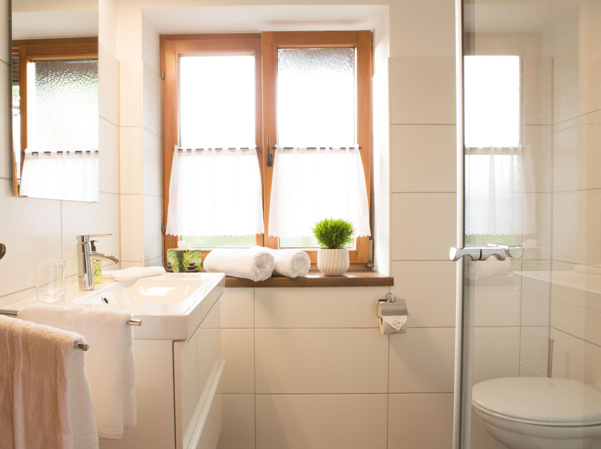 A bathroom with a sink, mirror, window with curtains, and a shower.
