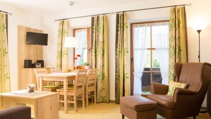 Una sala de estar acogedora con una mesa de comedor de madera, dos sillas, un sillón de cuero marrón y un pequeño otoman. Un televisor de pantalla plana está montado en la pared, y hay una lámpara sobre la mesa.