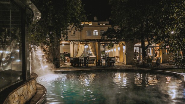Zona de piscina al aire libre con jacuzzi, rodeada de mesas y sillas, y un edificio con porche cubierto al fondo.