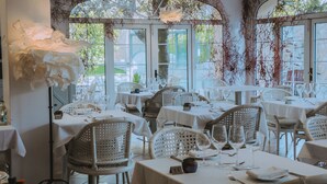 Un restaurante con mesas blancas y sillas de mimbre, amplias ventanas con vistas a la vegetación y enredaderas colgantes.