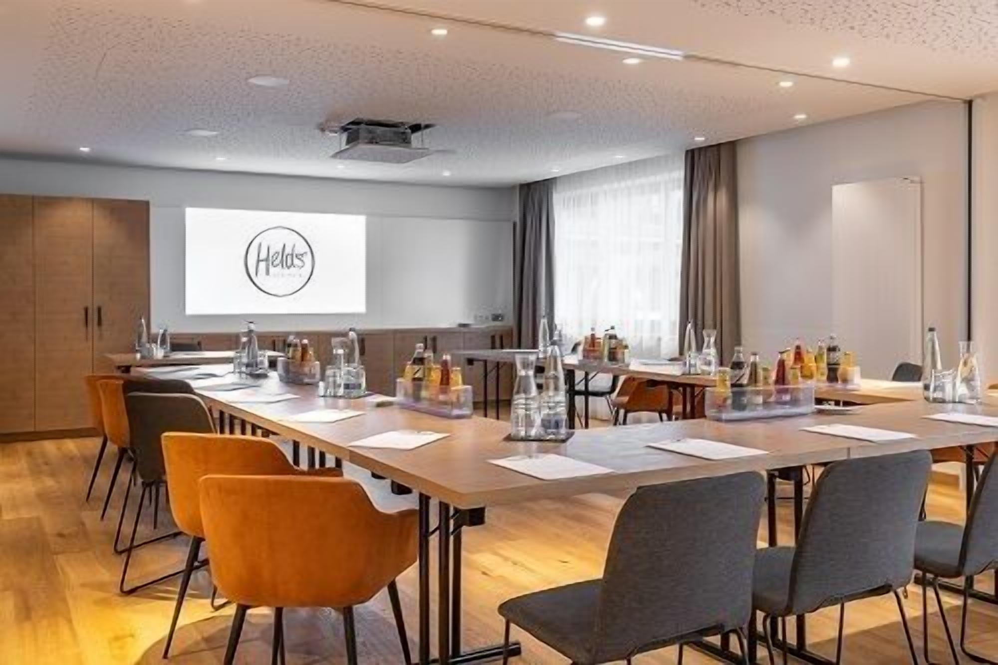 Une salle de conférence avec une longue table, des chaises et un écran de projection affichant le mot « Helds ».