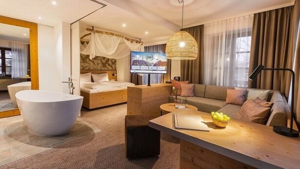 A modern hotel room with a large bathtub, a sofa, a TV, and a dining area with a laptop and fruit bowl. - Family room 30 to 40 sqm, sep. Children's bedroom, fridge, shower, WC (Ruhpolding)