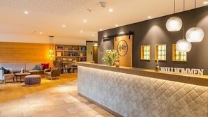 A hotel reception area with a modern design, featuring a reception desk, a seating area with sofas and a coffee table, and a decorative wall with framed pictures and a sign. - Family room 30 to 40 sqm, sep. Children's bedroom, fridge, shower, WC (Ruhpolding)