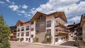 A multi-story hotel with a prominent logo, balconies, and a clear sky. - Family room 30 to 40 sqm, sep. Children's bedroom, fridge, shower, WC (Ruhpolding)
