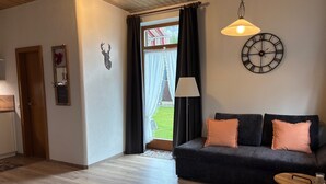 En stue med en mørk sofa, et rundt sofabord, et vægur og et vindue med gardiner.