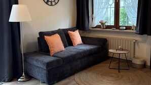 En moderne stue med en mørkegrå sofa, et rundt bord, en gulvlampe, et vægur og et vindue med lette gardiner.