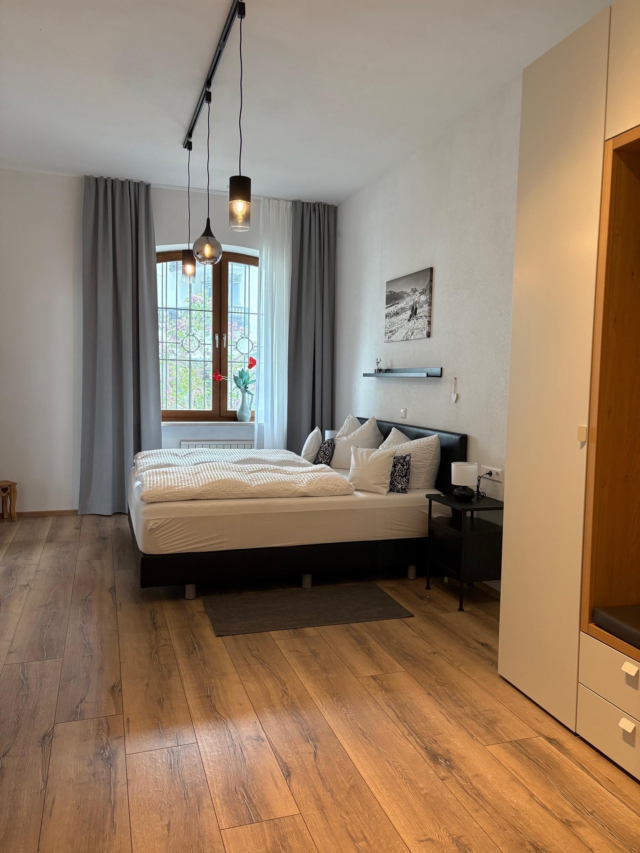 Ein Schlafzimmer mit Bett, Nachttisch, Lampe, einem Fenster mit Vorhängen und einem Bild an der Wand.