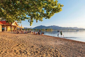 A beach with people relaxing under a tree, a wooden structure, and a lake with mountains in the background. - Vacation apartment for 2-4 persons, 80 sqm, 2 bedrooms, balcony (Marquartstein)