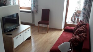 Ein kleiner, gut beleuchteter Raum mit Fernseher, einem roten Sofa, einem Sessel und einer Wanduhr.