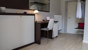 Une petite pièce moderne, équipée d’un bureau, d’une chaise et d’un miroir au-dessus du bureau. Un radiateur est fixé au mur, et un porte-vêtements est suspendu.