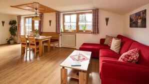 A cozy living room with a red sofa, wooden dining table, and chairs. There is a window with curtains, a radiator, and a painting on the wall. - Ferienwohnung Panoramablick 2 - 5 Personen (Berchtesgaden)