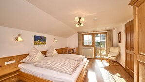 A bedroom with a wooden bed, a desk, a chair, and a view of the outdoors through a large window. - Ferienwohnung Panoramablick 2 - 5 Personen (Berchtesgaden)