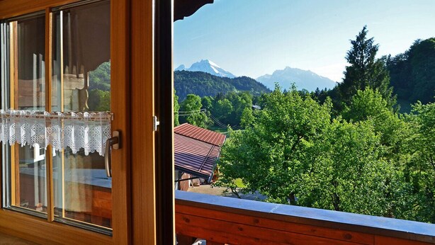 A wooden-framed window with a lace curtain, overlooking a mountainous landscape with trees and a house. - Ferienwohnung Panoramablick 2 - 5 Personen (Berchtesgaden)