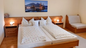 Un lit double avec une literie à motifs floraux, deux tables de chevet avec des lampes, une tête de lit en bois, une chaise et un tableau représentant un paysage de montagne accroché au mur.