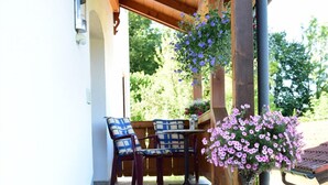 A covered outdoor seating area with two chairs and a table, surrounded by hanging flower baskets.