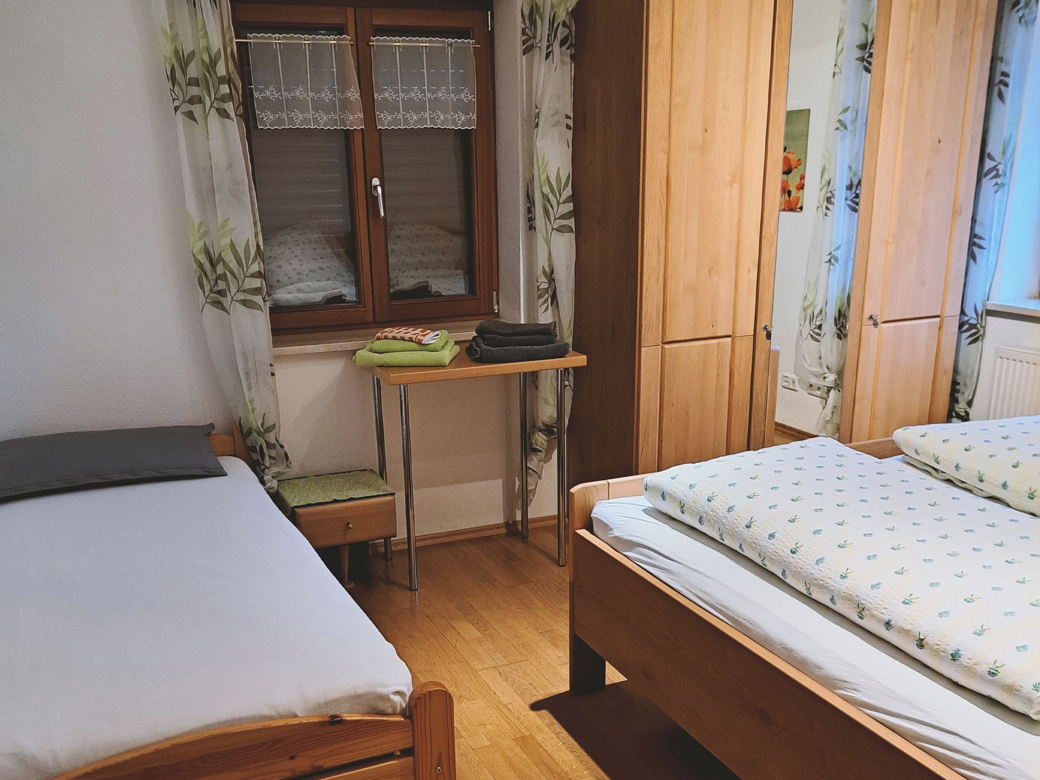 Un dormitorio con dos camas, un armario de madera, una mesita con toallas y una ventana con cortinas.