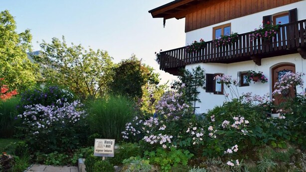 A traditional chalet with a wooden balcony, surrounded by lush greenery and flowering plants.