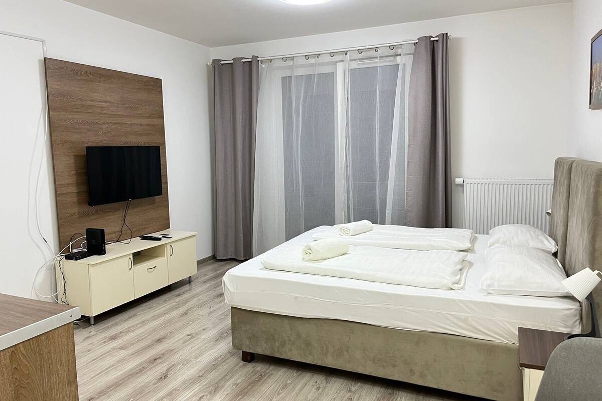 Habitación de hotel con cama, televisor montado en un panel de madera, escritorio con computadora y ventana con cortinas.