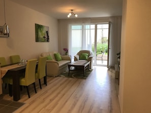 A modern living room with a dining area, a sofa, a coffee table, and a view of the outdoors through large windows. - Corvin Rose Apartment Hotel (Budapest)