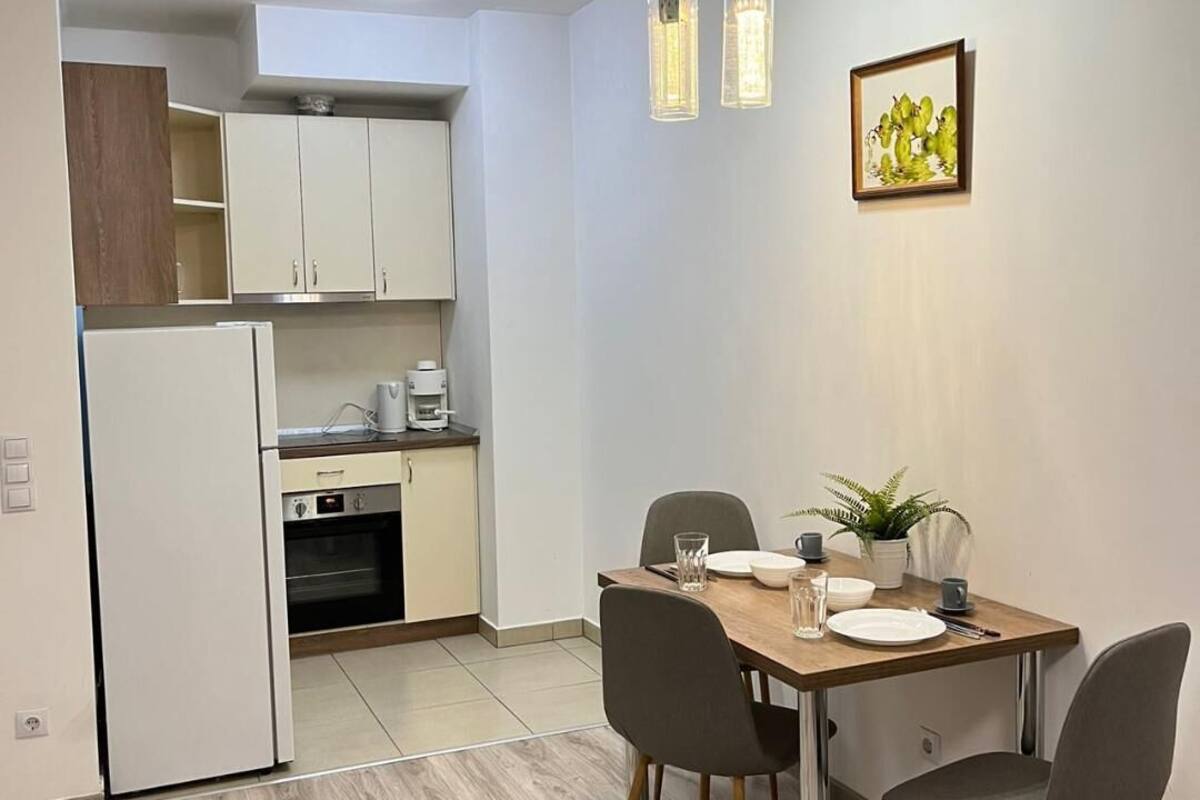 Una cocina moderna con armarios blancos, un refrigerador y una cafetera. Hay un comedor con una mesa de madera, cuatro sillas y una planta en maceta.