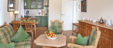 Un salon chaleureux avec un canapé à carreaux, des chaises en bois, une table en bois sur laquelle se trouve un panier de fruits, et une cuisine en arrière-plan.