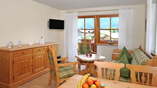 Ein gemütliches Zimmer mit Holztisch, Obstschale, Wandfernseher und einem fenster mit Vorhängen.