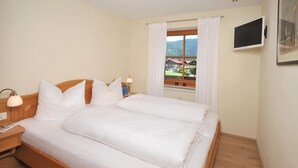 Ein Schlafzimmer mit einem hölzernen Bett, weißer Bettwäsche, einem Nachttisch mit Lampe, einem Fenster mit Vorhängen und einem an der Wand befestigten Fernseher.