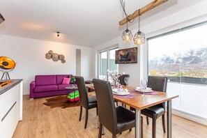 A modern dining area with a wooden table set for two, a purple sofa, and a city view through large windows. - Apartment 2 Deluxe Studio, 52 sqm, 1-4 persons, 1 bedroom, living room and balcony (Bayerisch Gmain)