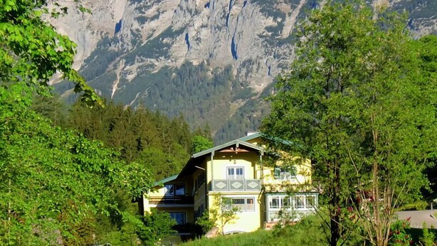 A two-story house with a balcony, surrounded by dense greenery and a mountain backdrop.