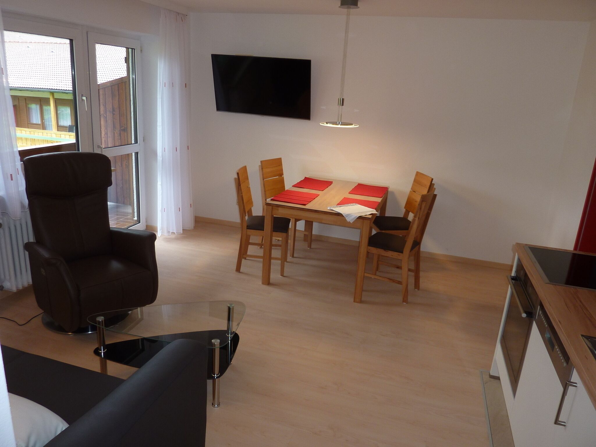 A modern living room with a dining area, a flat-screen TV mounted on the wall, and a large window with curtains.
