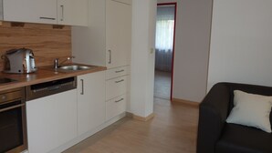 Cozinha moderna com armários brancos, uma pia de aço inoxidável e uma cafeteira. Há um sofá preto com uma almofada branca na sala de estar.